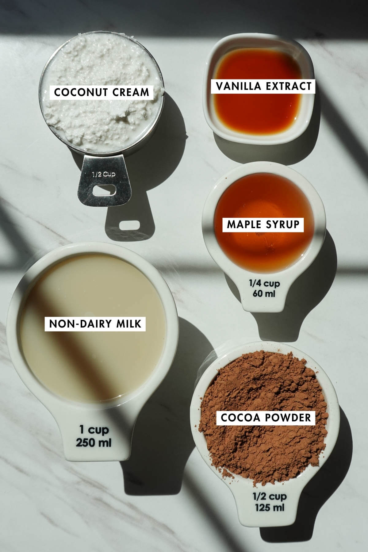 Ingredients for healthy fudgesicles on a table including coconut cream, cocoa powder, non-dairy milk, vanilla extract, and maple syrup.