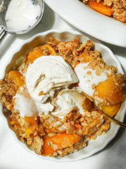 Ramekin with a serving of healthy peach crisp with vegan vanilla ice cream melting on top.
