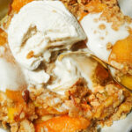 Spoon scooping warm peach crisp with nutty oat topping and melting vegan ice cream