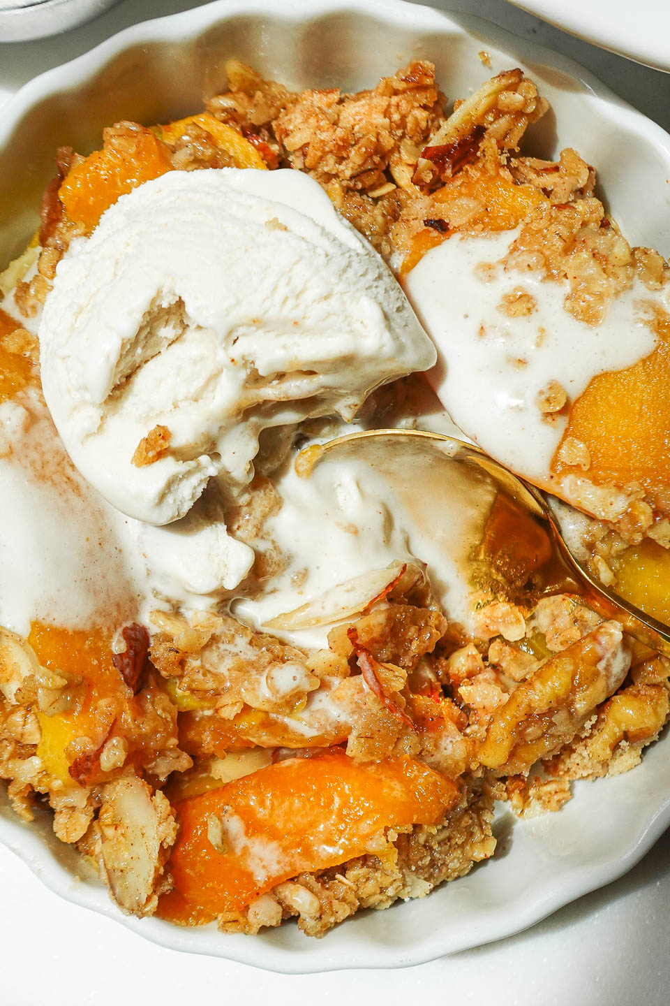 Spoon scooping warm peach crisp with nutty oat topping and melting vegan ice cream