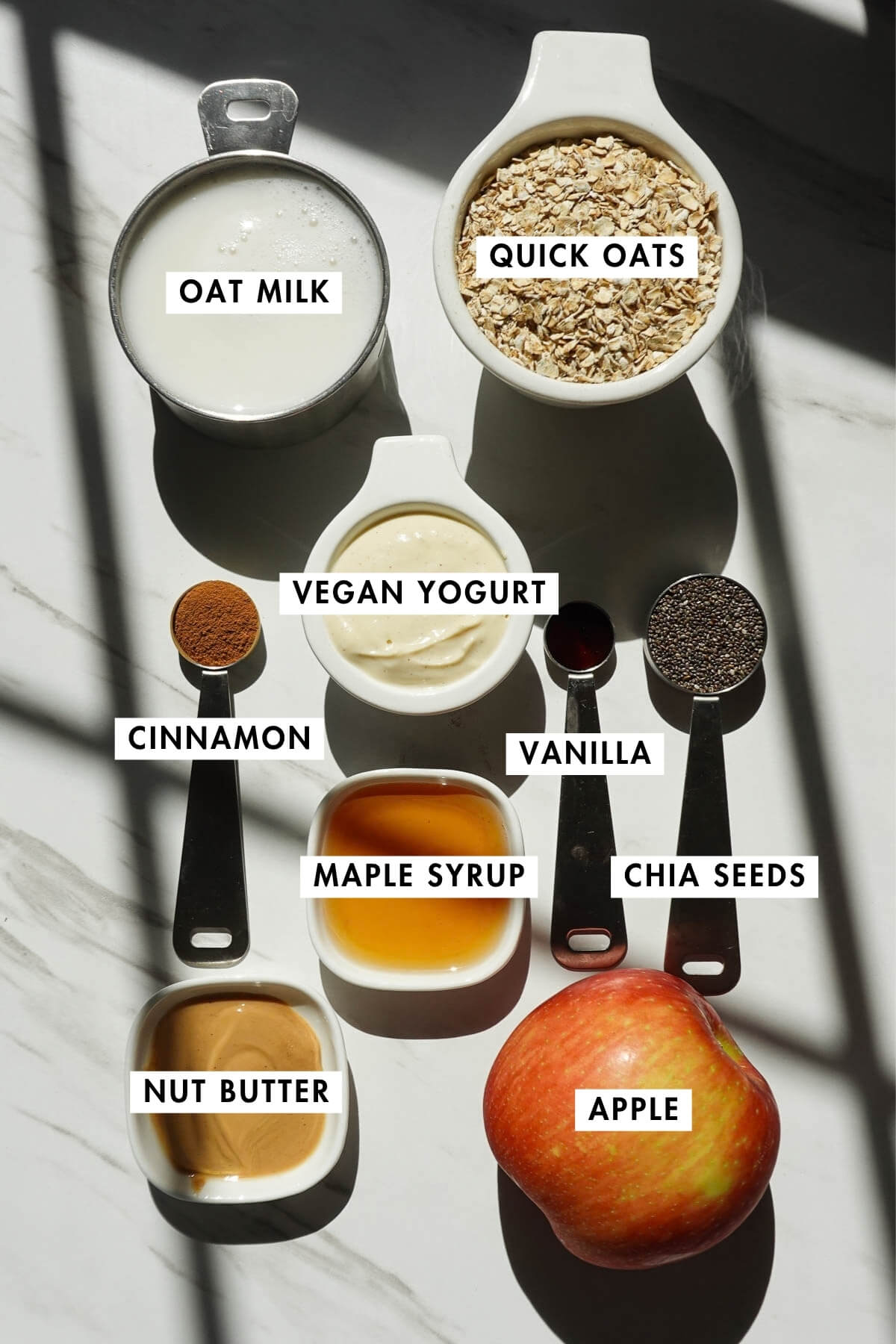Ingredients for apple cinnamon overnight oats including oats, oat milk, apple, vegan yogurt, nut butter, chia seeds, cinnamon, and maple syrup.