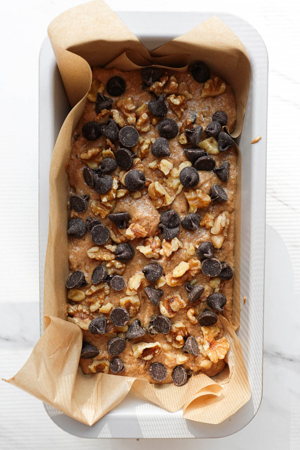 Banana bread in a loaf pan before baking, topped with chopped walnuts and chocolate chips.