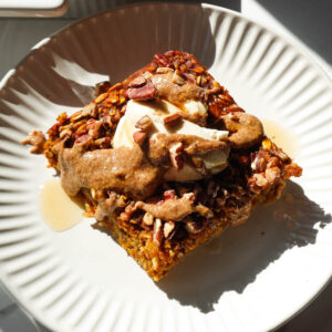 A slice of pumpkin baked oatmeal topped with vegan yogurt and a drizzle of maple syrup and almond butter.