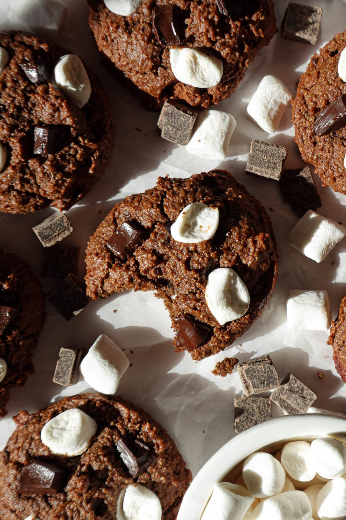Overhead shot of vegan hot chocolate cookies with a bite taken out arranged on parchment paper, topped with chocolate chips and mini marshmallows, perfect for holiday baking.