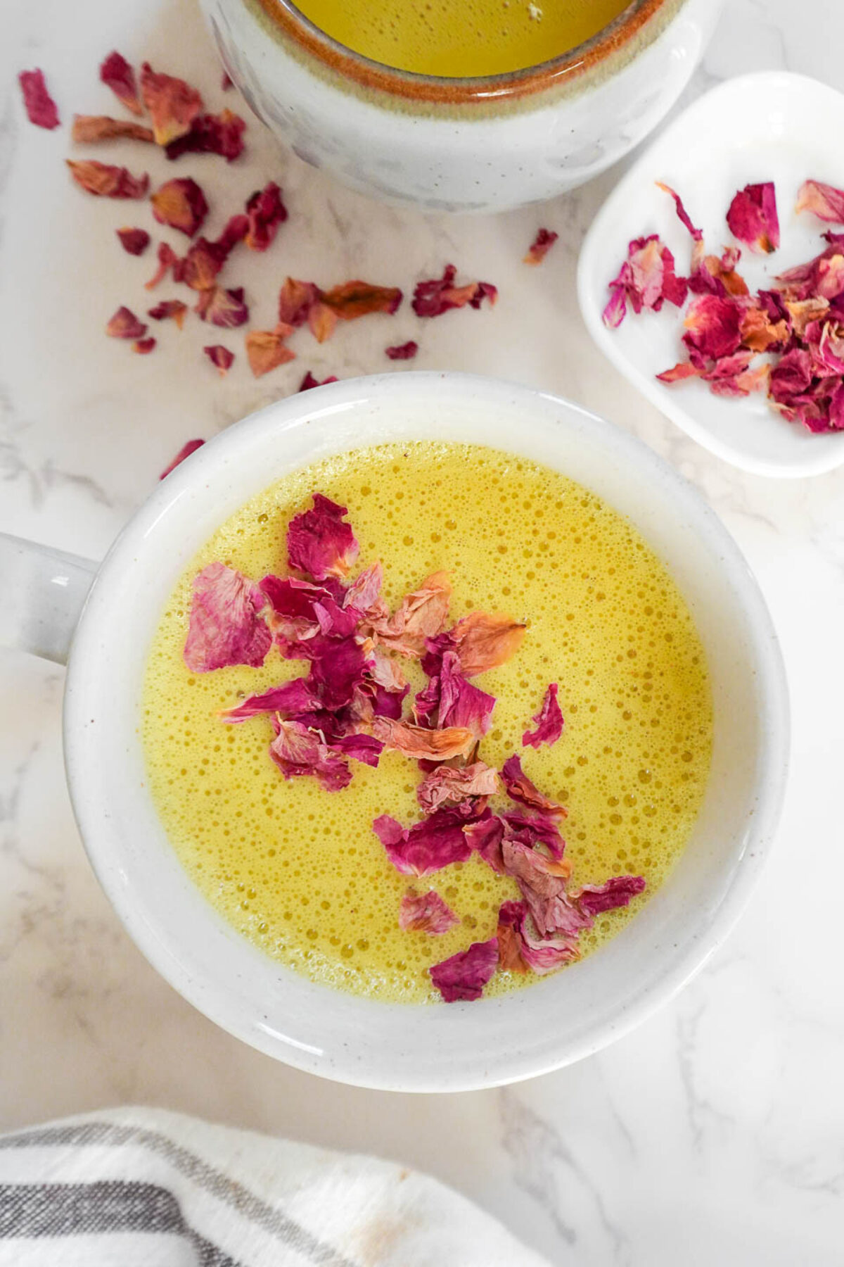 A cup of golden milk (turmeric latte) with a frothy, creamy texture, garnished with rose petals on a marble surface.