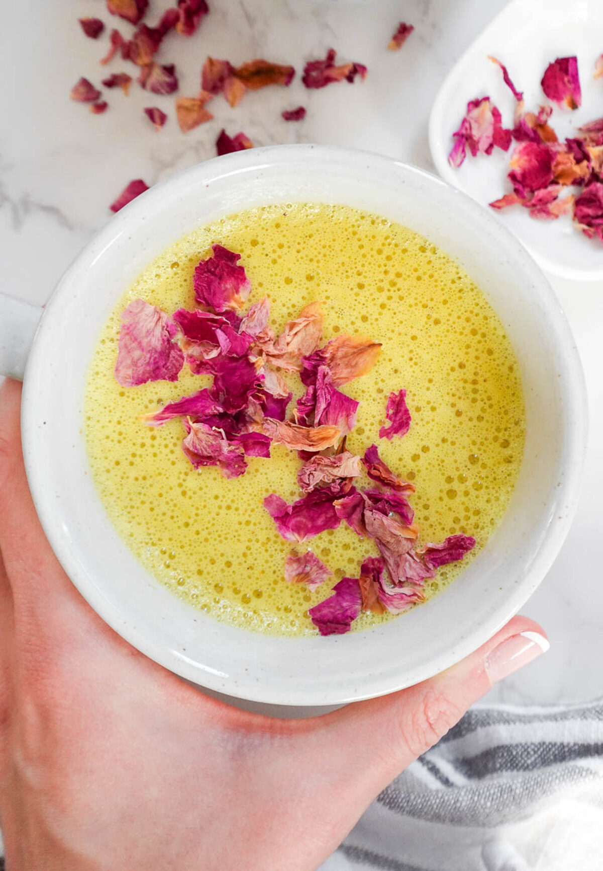 A cup of golden milk (turmeric latte) with a frothy, creamy texture, garnished with rose petals on a marble surface.