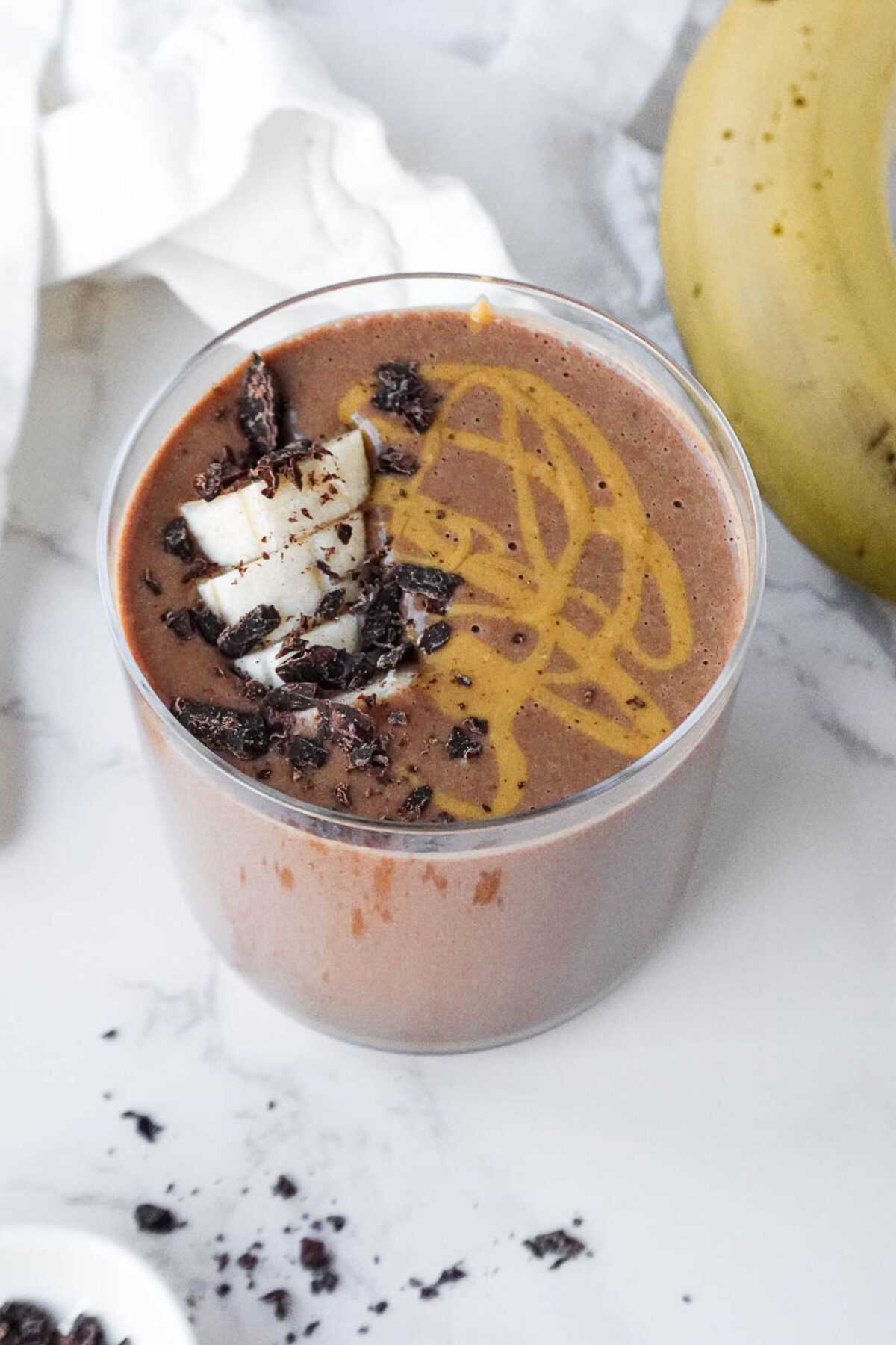 vegan chocolate banana smoothie served in a glass topped with cacao nibs, sliced banana, and a drizzle of peanut butter