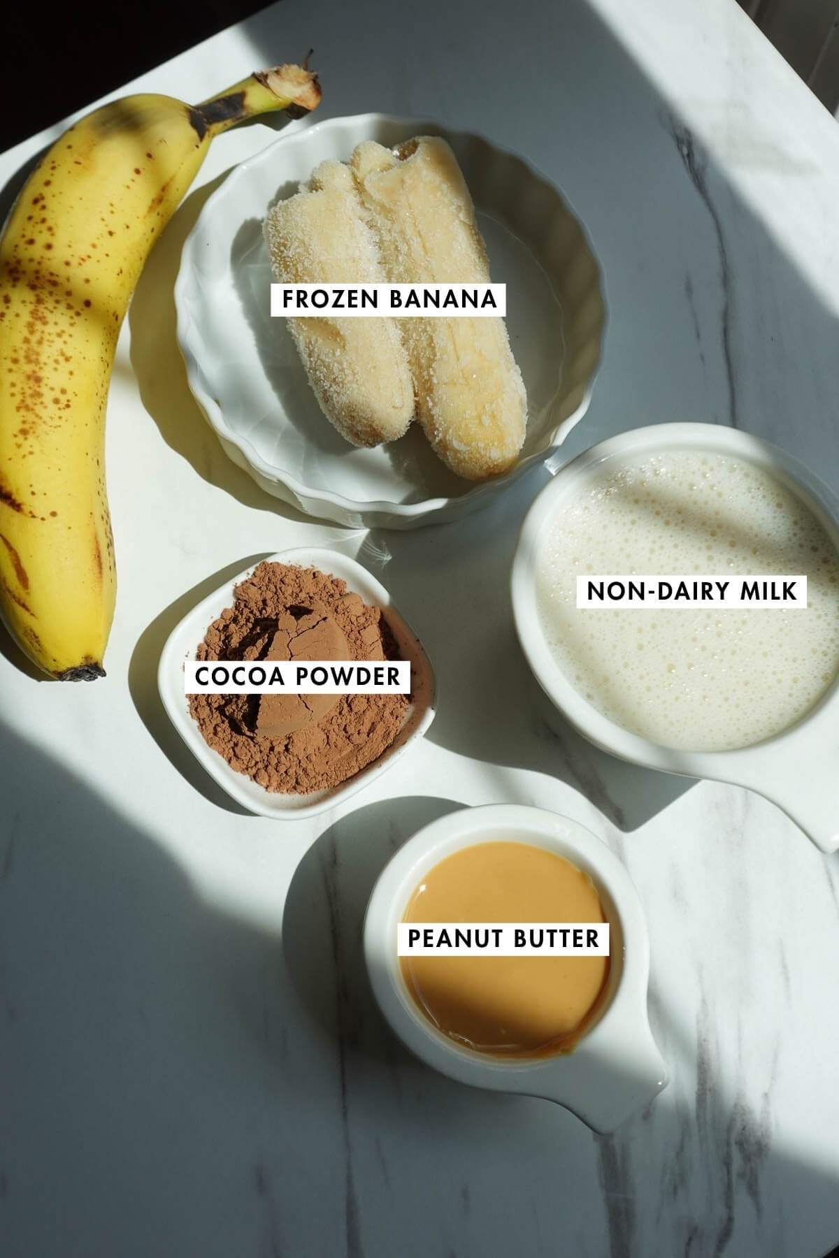 Ingredients for a chocolate peanut butter banana smoothie on a table including frozen banana, cocoa powder, peanut butter, and non-dairy milk.