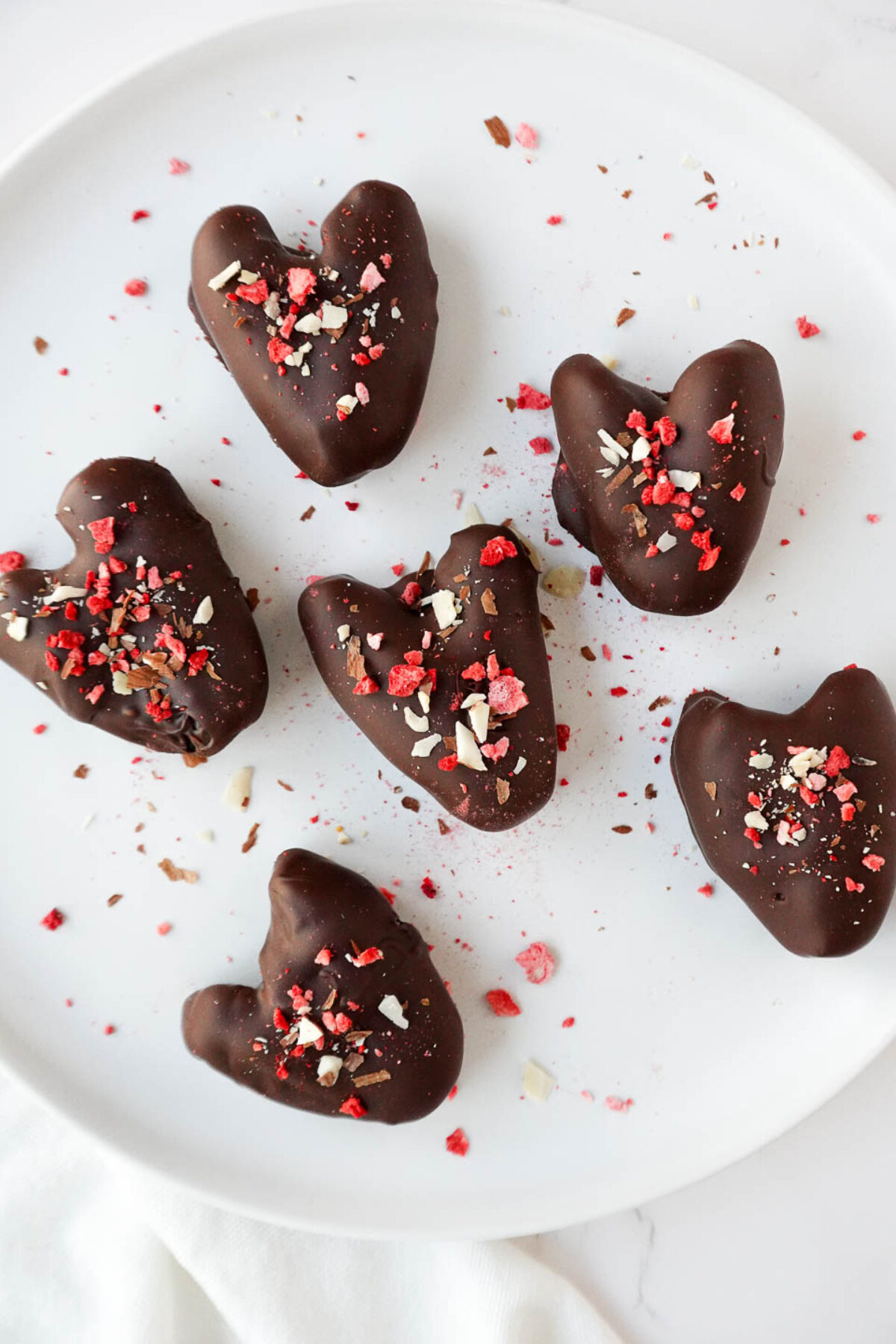 Heart shaped chocolate covered dates on a white plate topped with freeze-dried strawberries, sliced almonds, and flaky sea salt.