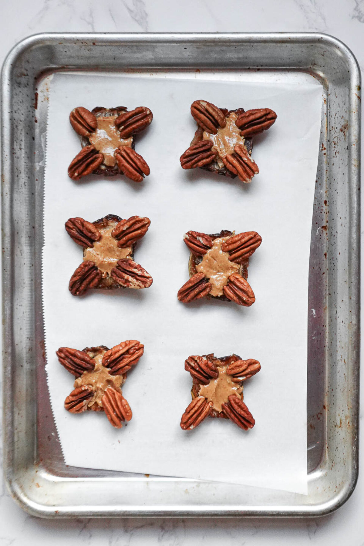 Medjool dates on a parchment-lined tray, topped with peanut butter and pecan halves to assemble chocolate turtles