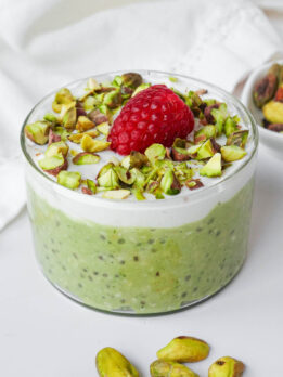 Vegan pistachio overnight oats in a glass jar topped with vegan yogurt, chopped pistachios, and one raspberry.