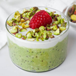 Vegan pistachio overnight oats in a glass jar topped with vegan yogurt, chopped pistachios, and one raspberry.