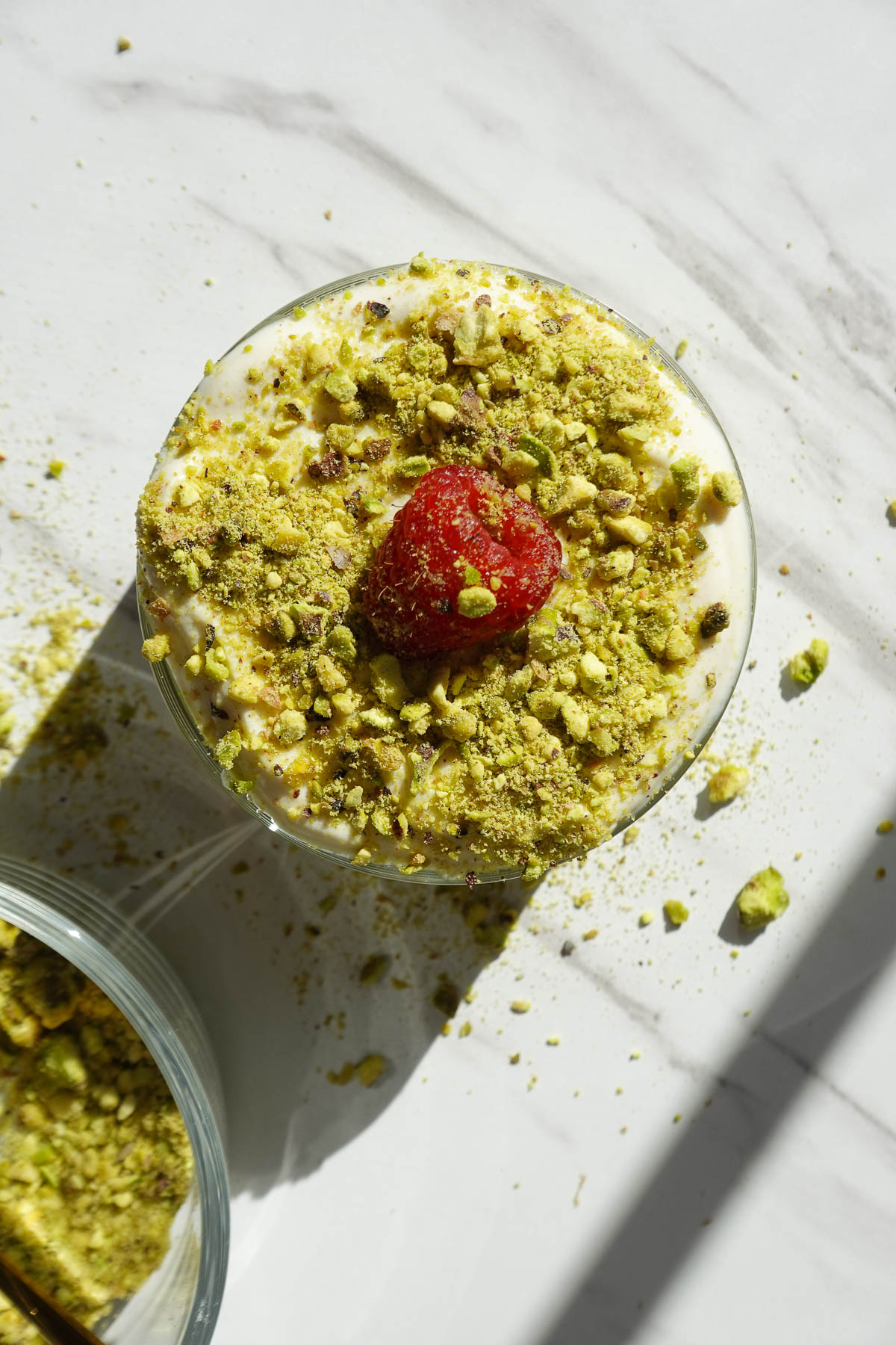 Overhead shot of vegan pistachio overnight oats in a glass jar topped with vegan yogurt, chopped pistachios, and one raspberry.