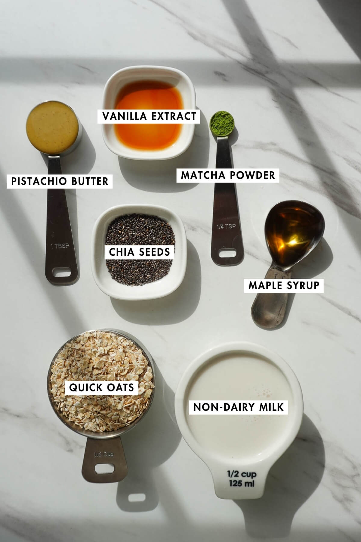 Measured ingredients needed to make pistachio overnight oats on a table including oats, chia seeds, non-dairy milk, maple syrup, matcha powder, vanilla extract, and pistachio butter.