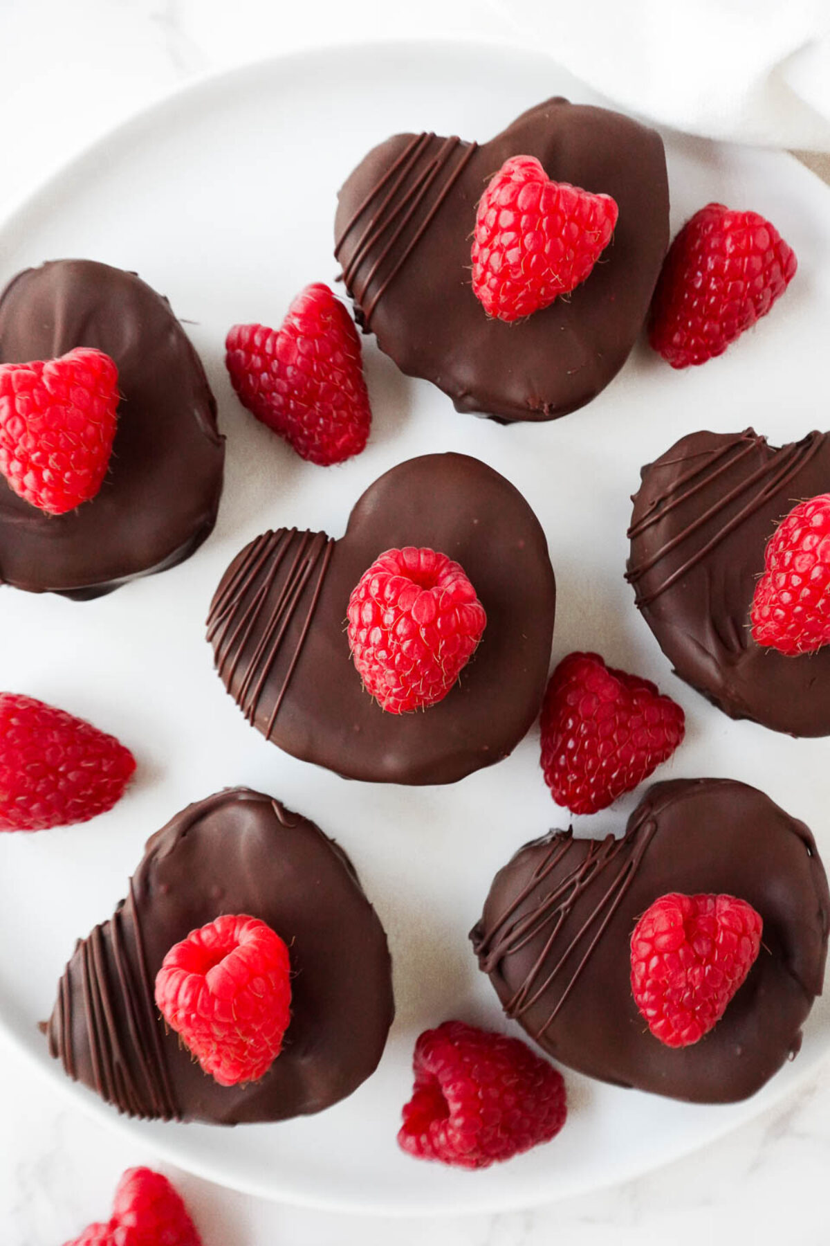 Overhead view of heart-shaped brownies on a white plate, drizzled with chocolate and garnished with fresh raspberries.