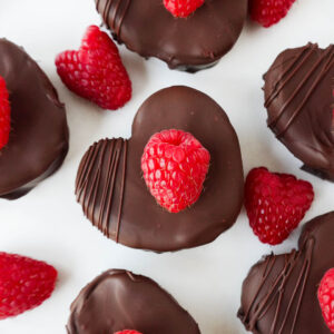Heart-shaped no-bake raspberry brownies coated in dark chocolate, garnished with a fresh raspberry on top.