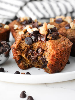 Moist and tender healthy banana muffins on a plate with a bite taken out showing gooey chocolate chips and it's fluffy, moist texture