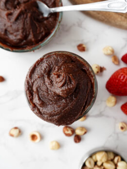 overhead shot of creamy homemade healthy Nutella in a glass jar, surrounded by hazelnuts and fresh strawberries, vegan and naturally sweetened with dates