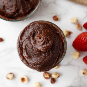overhead shot of creamy homemade healthy Nutella in a glass jar, surrounded by hazelnuts and fresh strawberries, vegan and naturally sweetened with dates