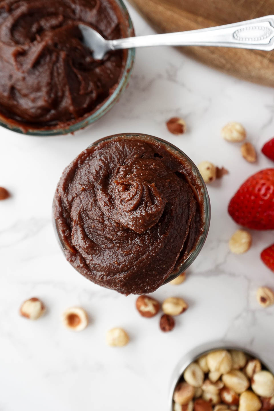 overhead shot of creamy homemade healthy Nutella in a glass jar, surrounded by hazelnuts and fresh strawberries, vegan and naturally sweetened with dates