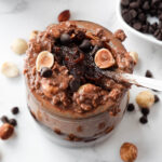 A spoon scooping up thick and creamy vegan chocolate hazelnut overnight oats with dark chocolate chips and hazelnuts on top and a spoonful of homemade healthy nutella