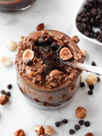A spoon scooping up thick and creamy vegan chocolate hazelnut overnight oats with dark chocolate chips and hazelnuts on top and a spoonful of homemade healthy nutella