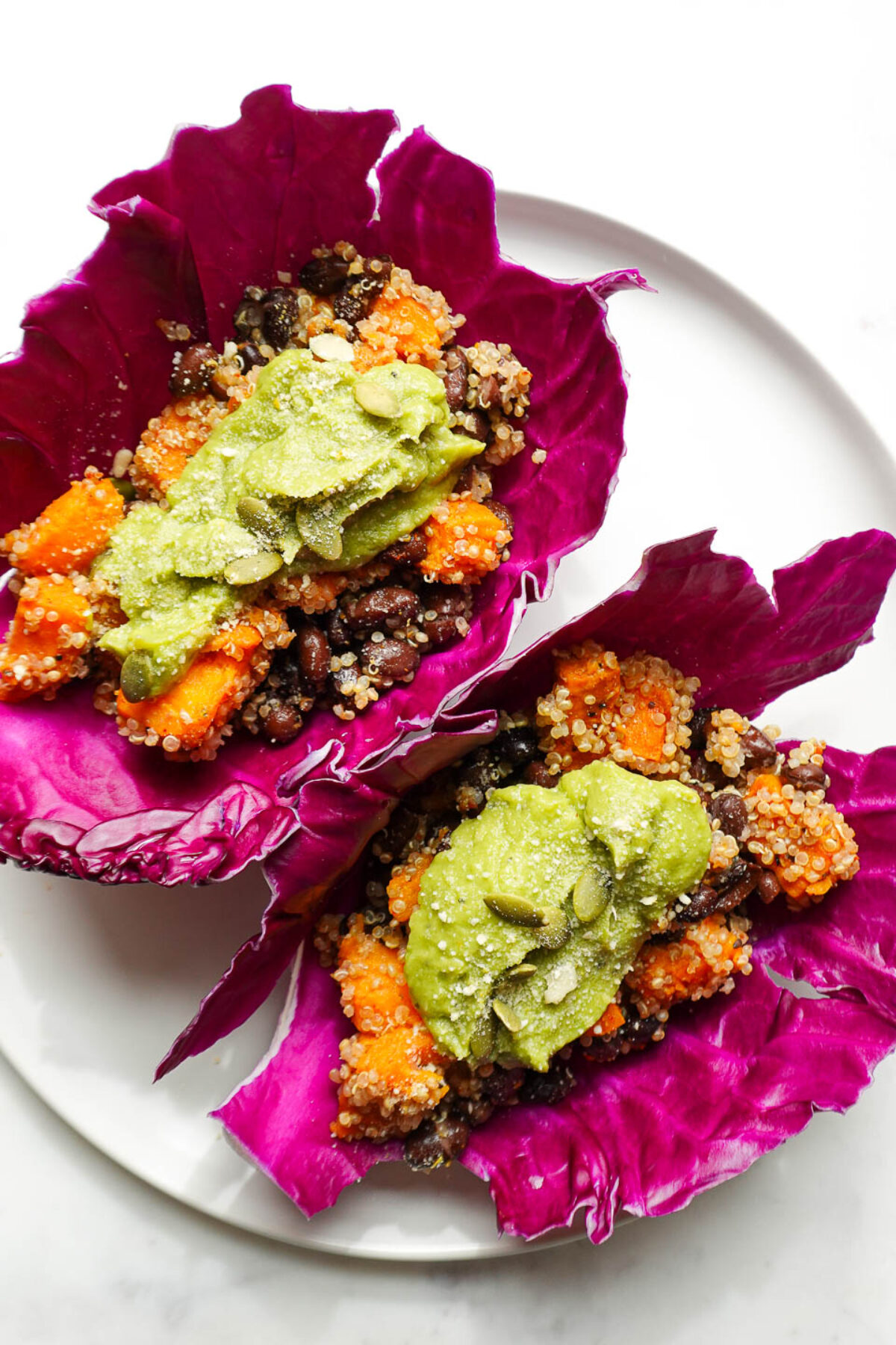 A colorful plate of two vegan sweet potato and black bean tacos in red cabbage wraps, topped with fresh guacamole.