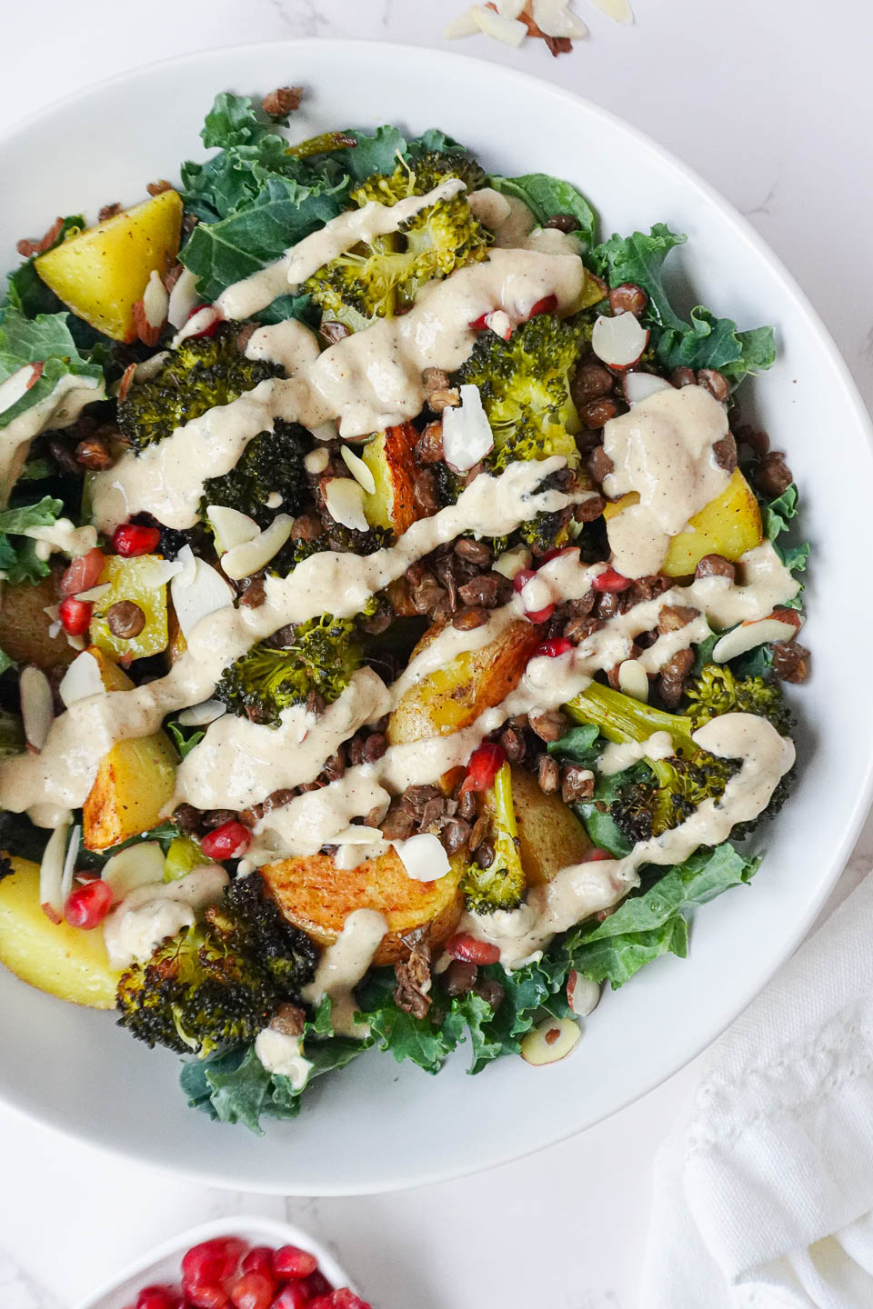 Roasted Potato & Lentil Buddha Bowl with crispy lentils, roasted broccoli, golden potatoes, sautéed kale, and topped with sliced almonds, pomegranate seeds, and a drizzle of creamy lemon tahini dressing.