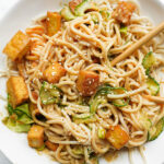 Bowl of peanut noodles topped with crispy tofu, spiralized cucumber, scallions, and sesame seeds with chopsticks