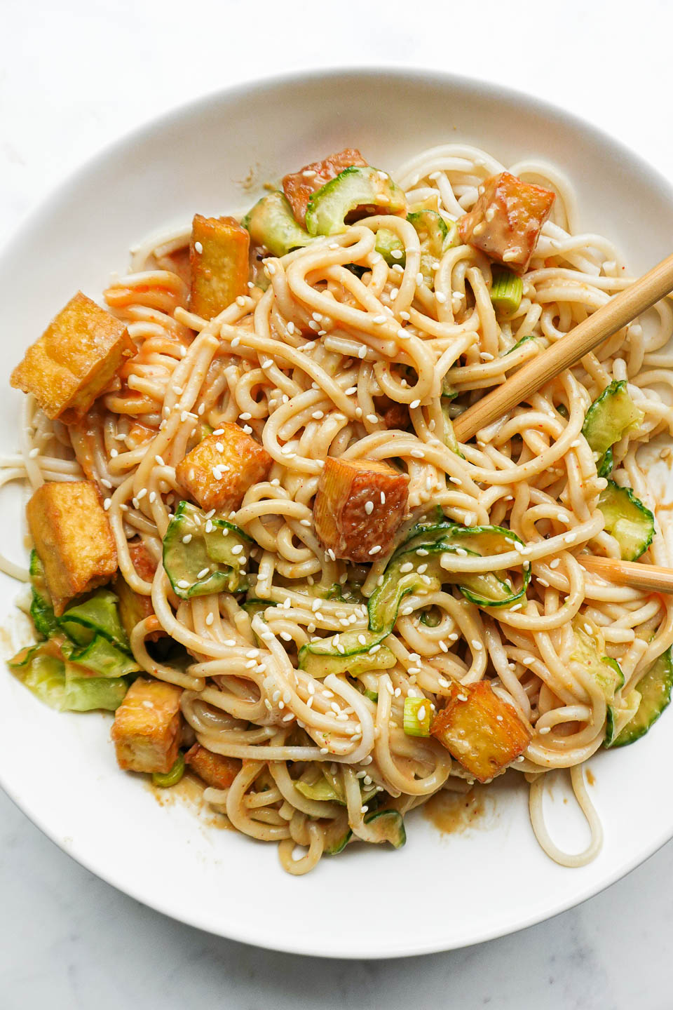 Bowl of peanut noodles topped with crispy tofu, spiralized cucumber, scallions, and sesame seeds with chopsticks
