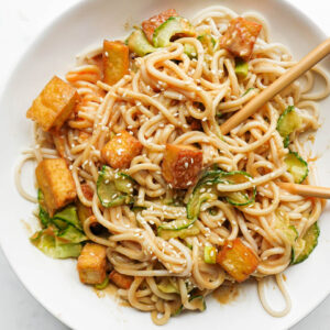 Healthy peanut noodles topped with crispy tofu, spiralized cucumber, scallions, and sesame seeds with chopsticks