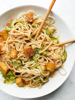 Bowl of peanut noodles topped with crispy tofu, spiralized cucumber, scallions, and sesame seeds with chopsticks
