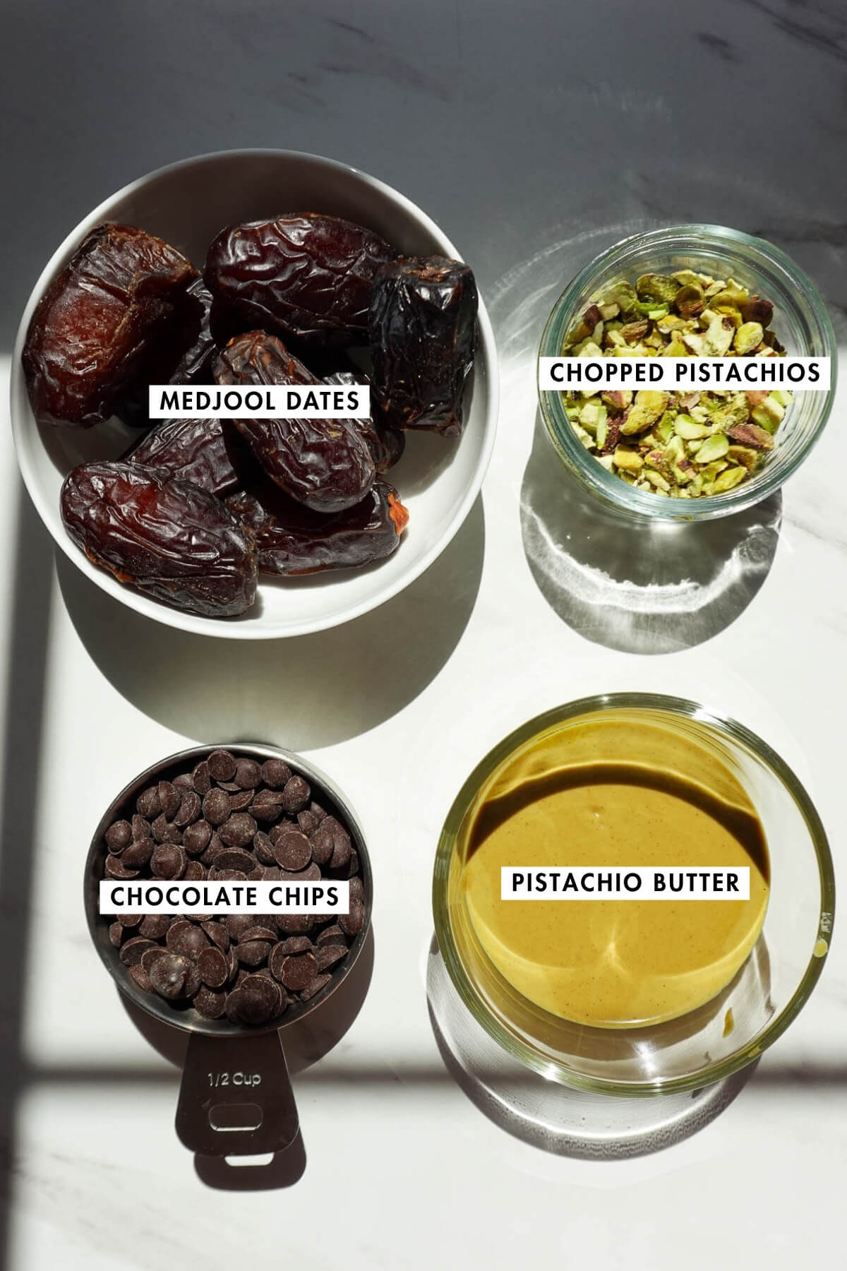 Ingredients needed to make pistachio stuffed dates on a table, including medjool dates, pistachio butter, chocolate chips, and chopped pistachios.
