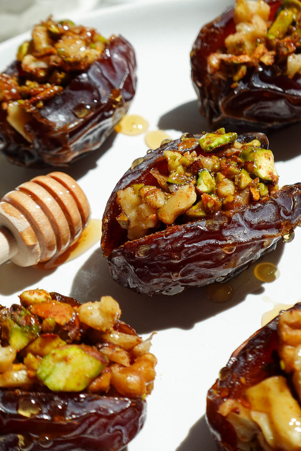 Baklava dates filled with pistachios, walnuts, and cinnamon arranged on a white plate with a honey comb.