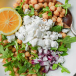 Overhead view of lemon chickpea salad with chickpeas, vegan feta, arugula, red onion, and lemon wedge.