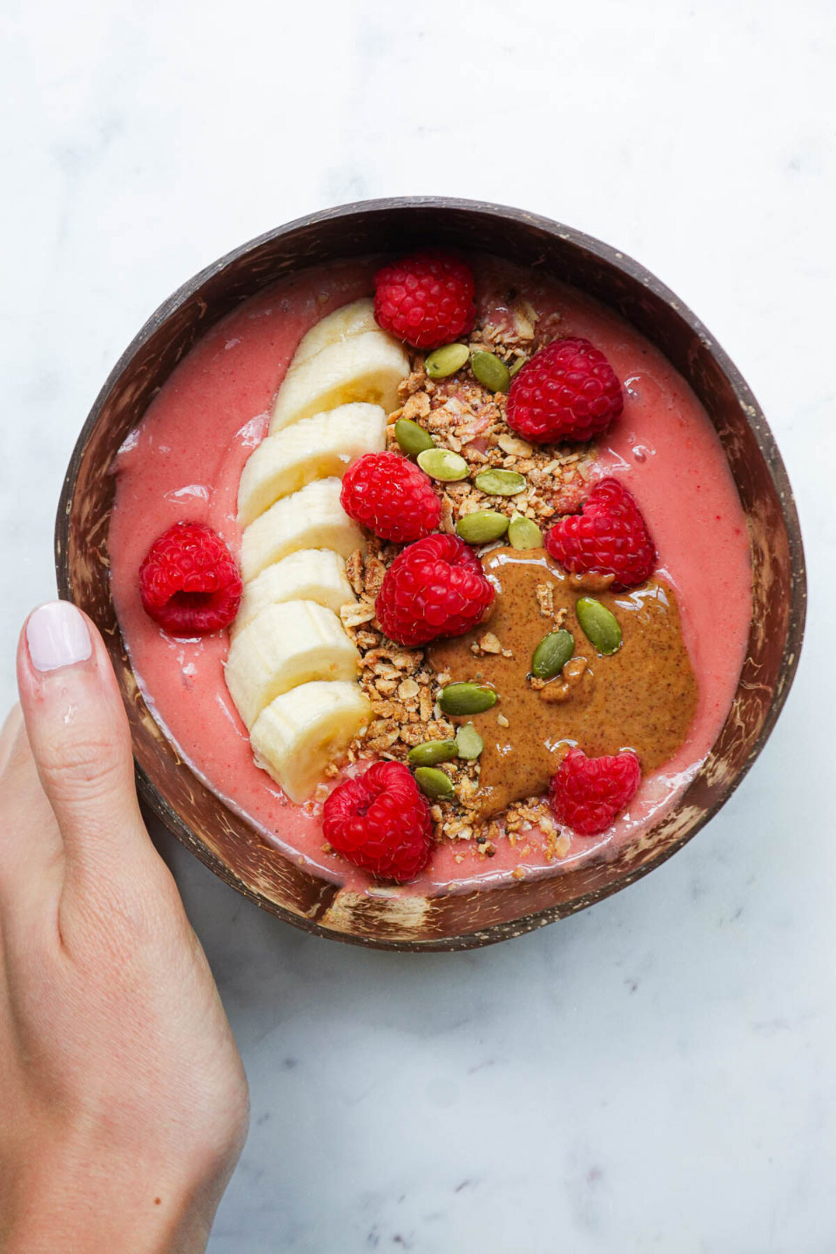 hand holding a vegan raspberry smoothie bowl in a coconut bowl topped with granola, sliced banana, fresh raspberries, almond butter, pumpkin seeds