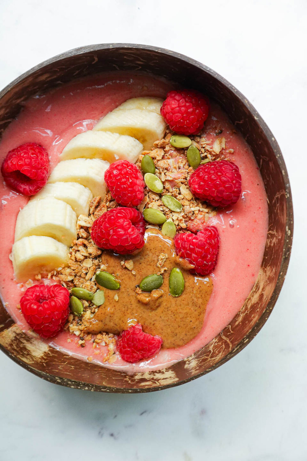 Close-up of raspberry smoothie bowl topped with granola, sliced banana, fresh raspberries, almond butter, pumpkin seeds