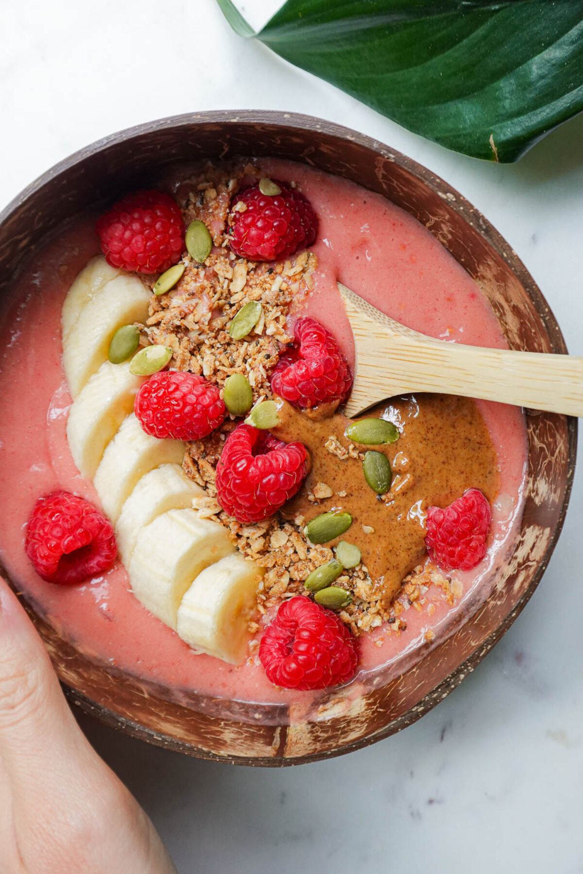 hand holding a vegan raspberry smoothie bowl with a spoon inside, in a coconut bowl, topped with granola, sliced banana, fresh raspberries, almond butter, pumpkin seeds