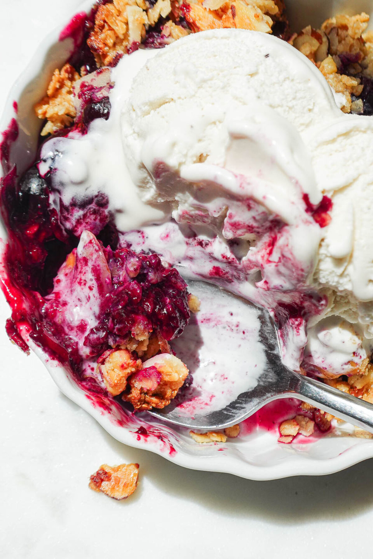 Close-up of crunchy crisp topping with oats and almonds over bubbling blueberries and swirls of vanilla ice cream.