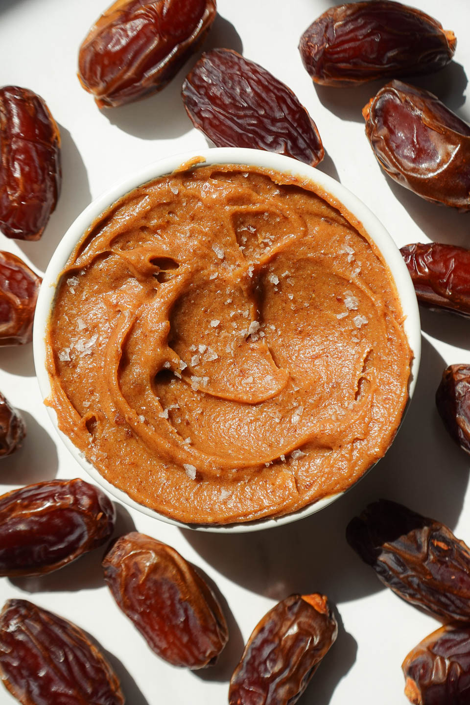 Healthy date caramel in a bowl surrounded by fresh dates and topped with flaky sea salt.