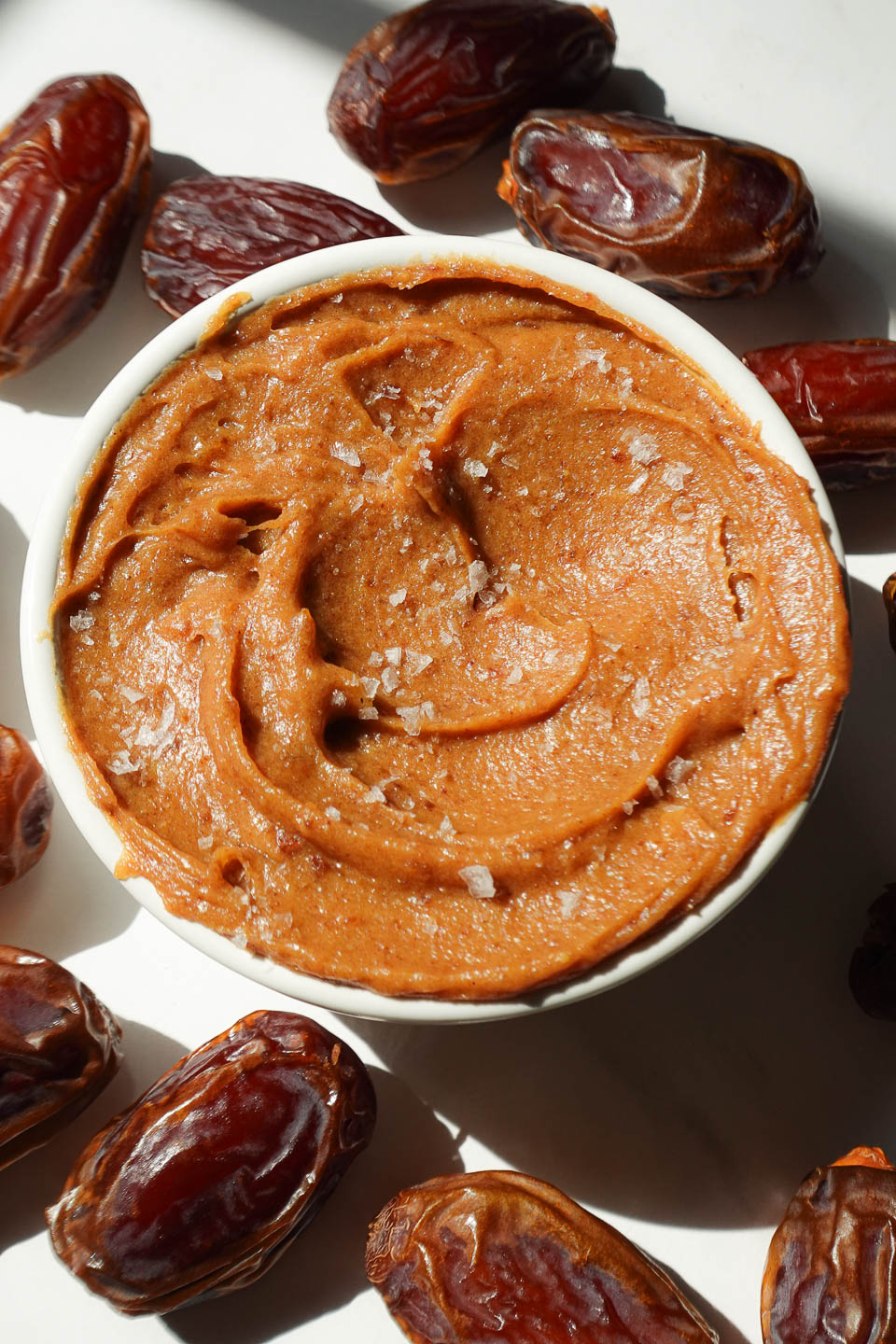 Healthy date caramel in a bowl surrounded by fresh dates and topped with flaky sea salt.