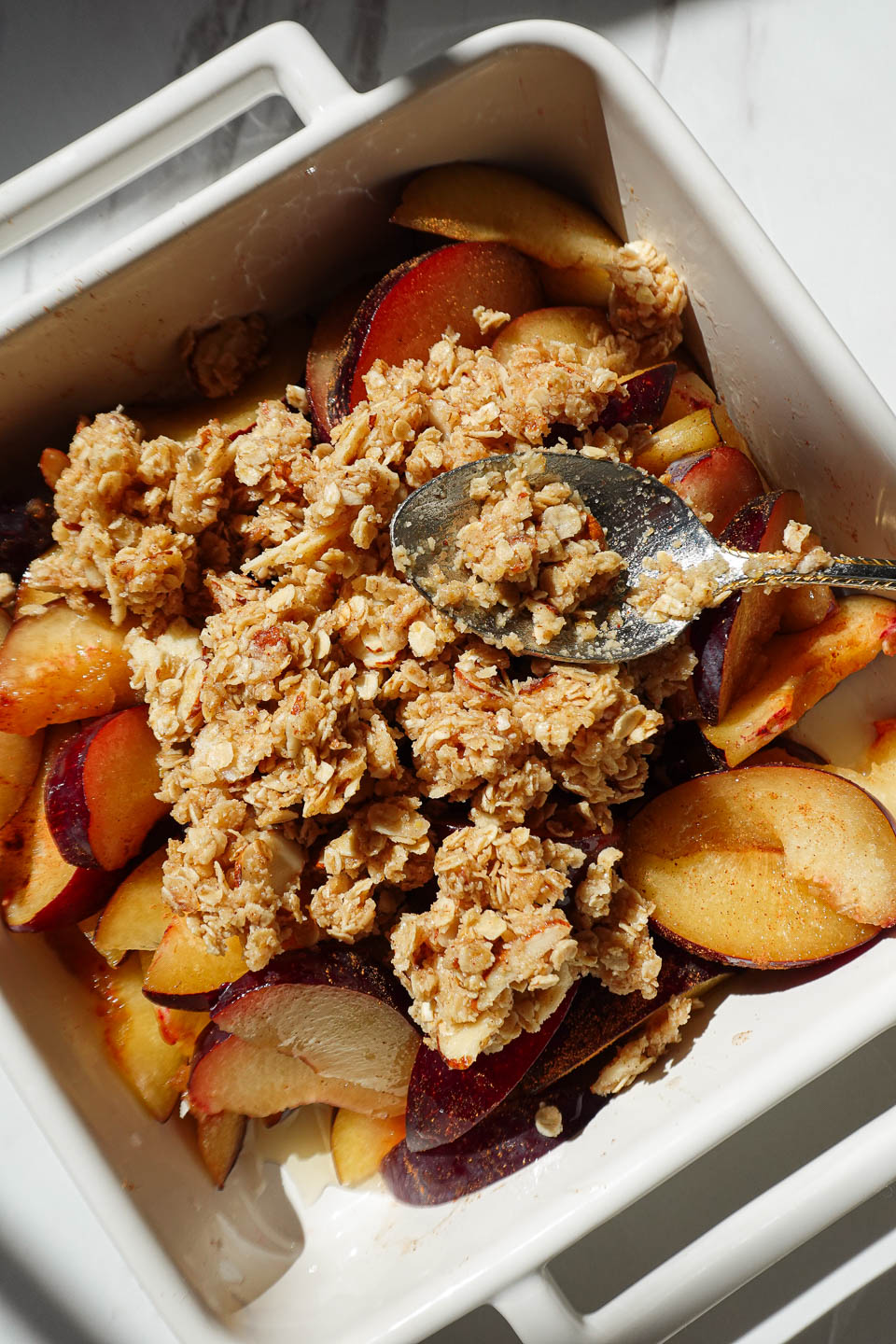 Almond oat crumble being sprinkled over sliced plums in a baking dish before baking