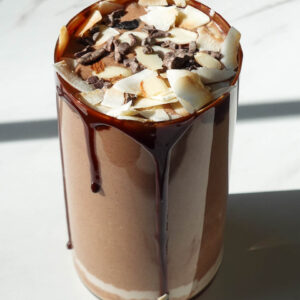 almond joy smoothie in a glass topped with coconut flakes, cacao nibs, and chocolate syrup