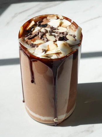 almond joy smoothie in a glass topped with coconut flakes, cacao nibs, and chocolate syrup