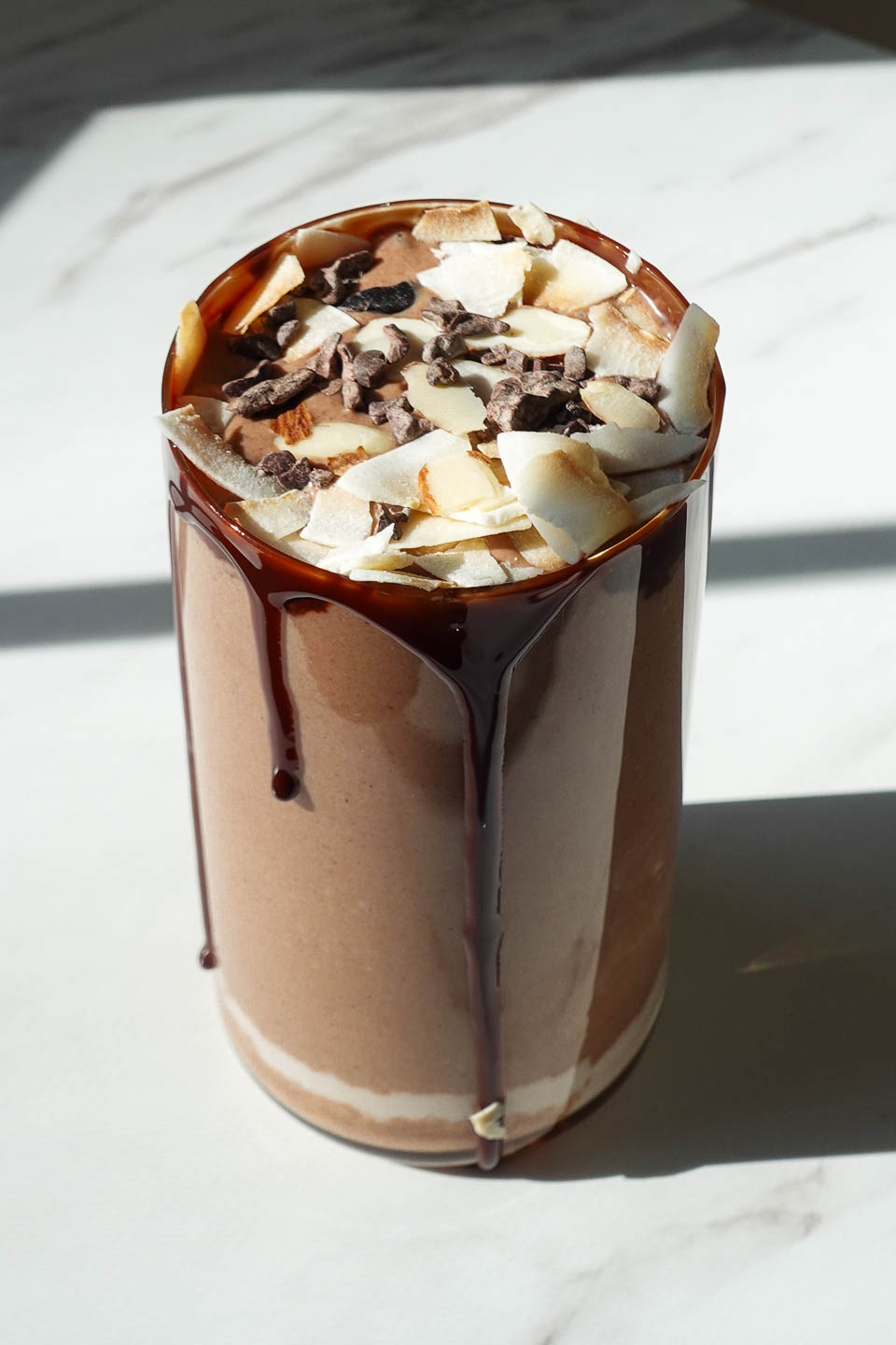 almond joy smoothie in a glass topped with coconut flakes, cacao nibs, and chocolate syrup