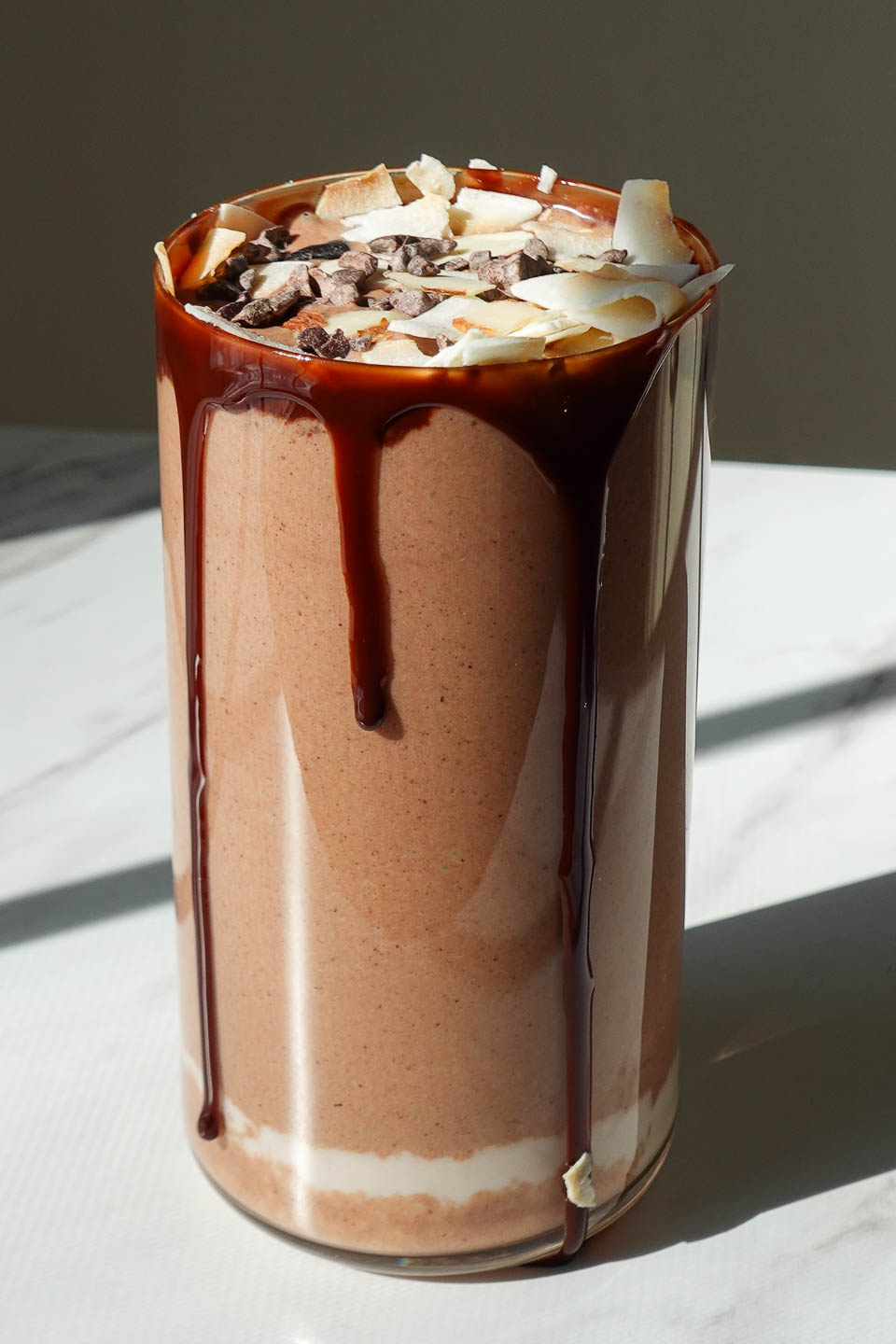 almond joy smoothie in a glass topped with coconut flakes, cacao nibs, and chocolate syrup