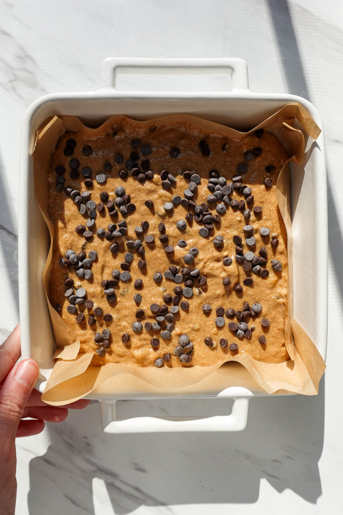 Mixture for vegan banana bars in a parchment-lined square baking pan with mini chocolate chips sprinkled on top.