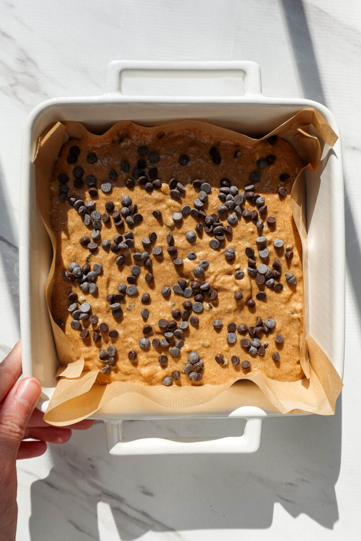 Vegan banana bars batter spread in a parchment-lined square baking pan with mini chocolate chips on top.