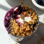 Blueberry muffin overnight oats in a glass jar topped with vegan yogurt, blueberry compote, and crushed protein cereal