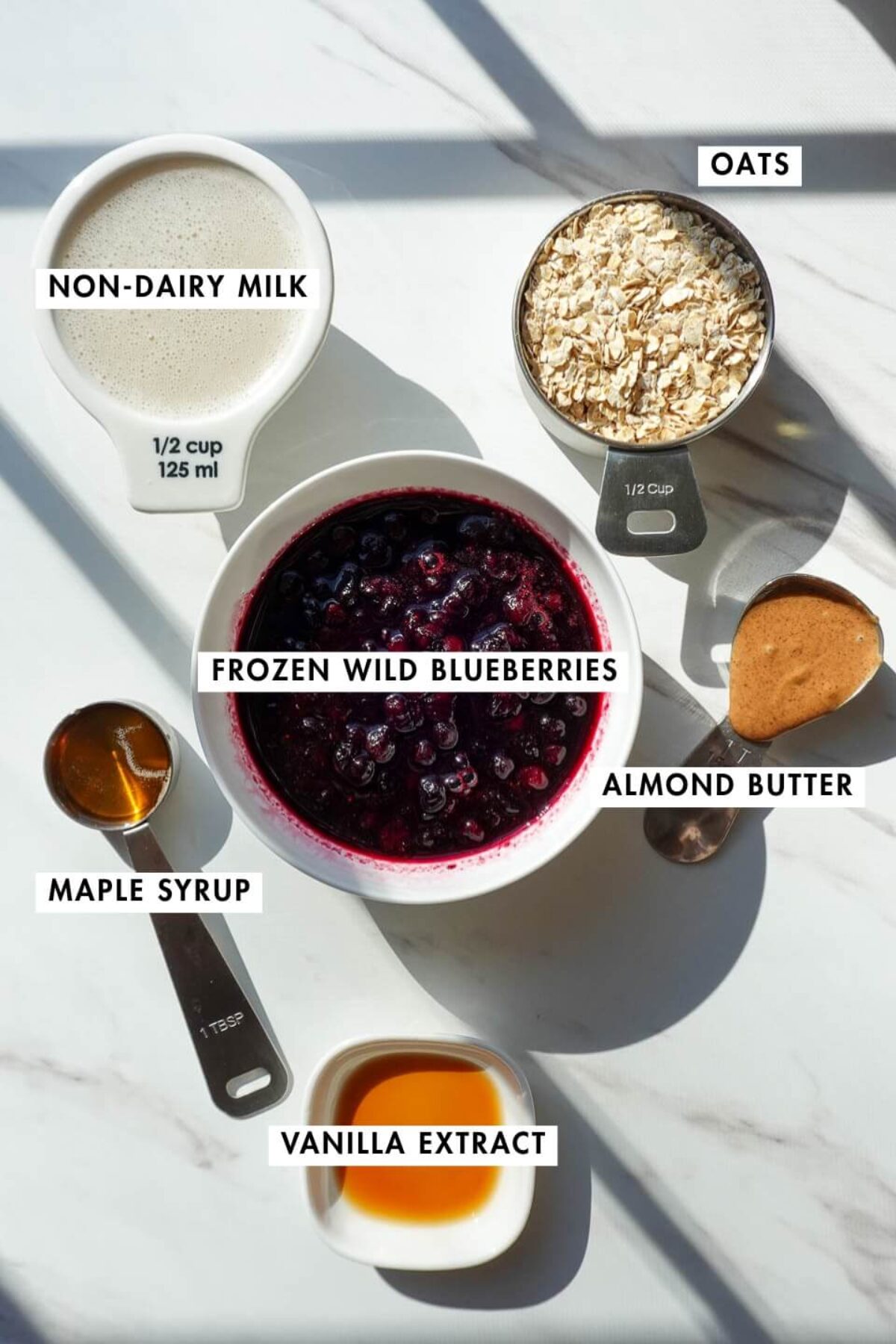 ingredients needed for blueberry muffin overnight oats including oats, frozen wild blueberries, almond butter, maple syrup, vanilla extract, and non-dairy milk on a countertop